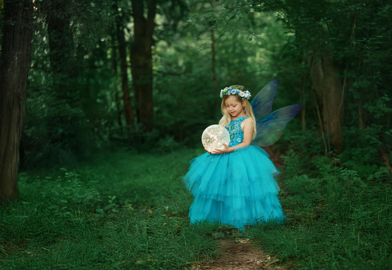 Fairy in a flowing blue gown holding a glowing lantern in a dreamy outdoor forest scene
