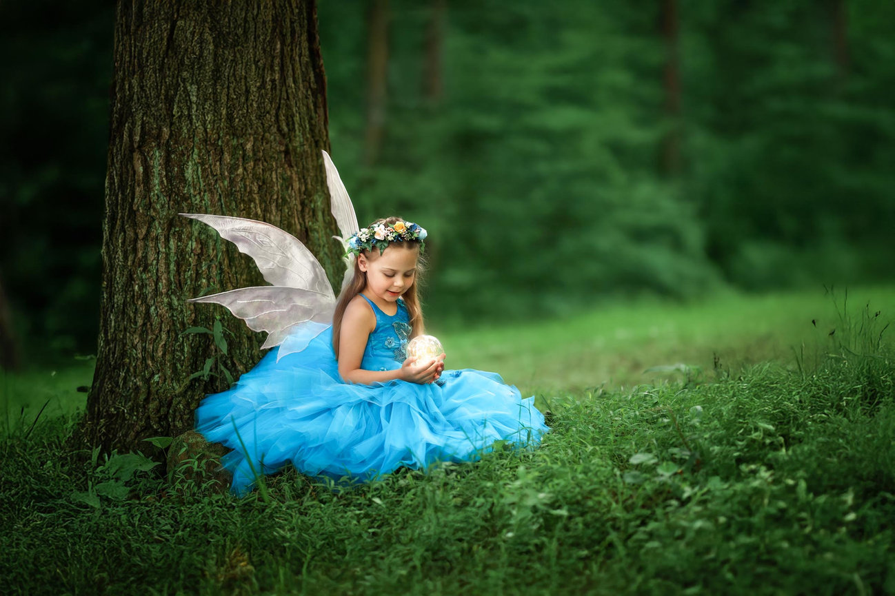 Fairy in a blue gown sitting at the foot of a tree surrounded by enchanted forest greenery