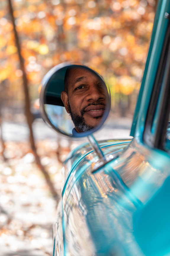 Reflection of a smiling man in a side mirror of a turquoise car, with blurred autumn leaves in the background in the style of Jo Hayes Images Charlotte portrait photography
