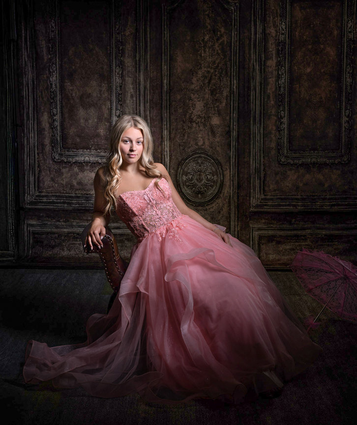 Portrait of a female high school graduate in her pink ballgown posing on a chair against a dark ornate studio backdrop