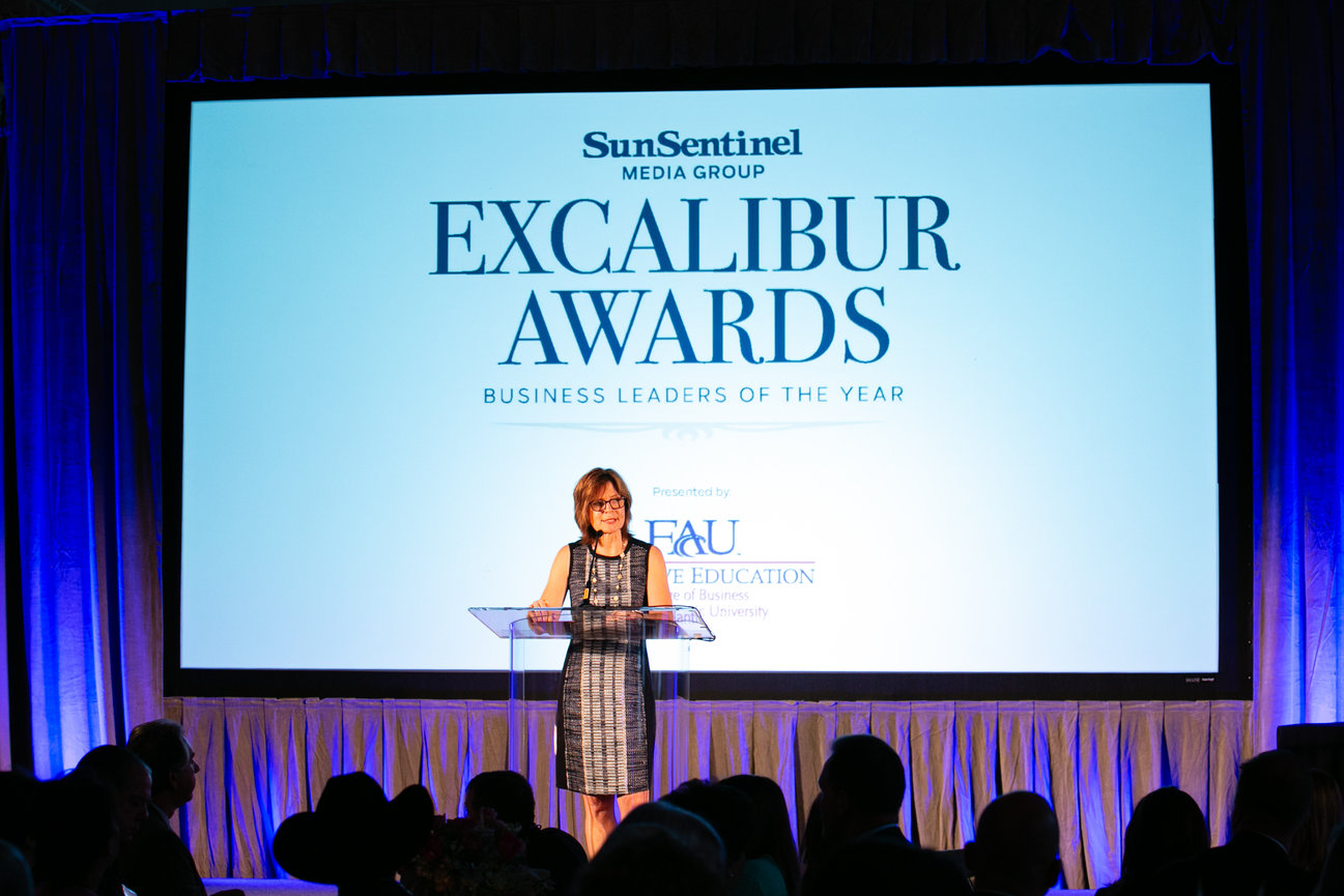Woman speaking at a podium during the Sun Sentinel Excalibur Awards ceremony on a lit stage