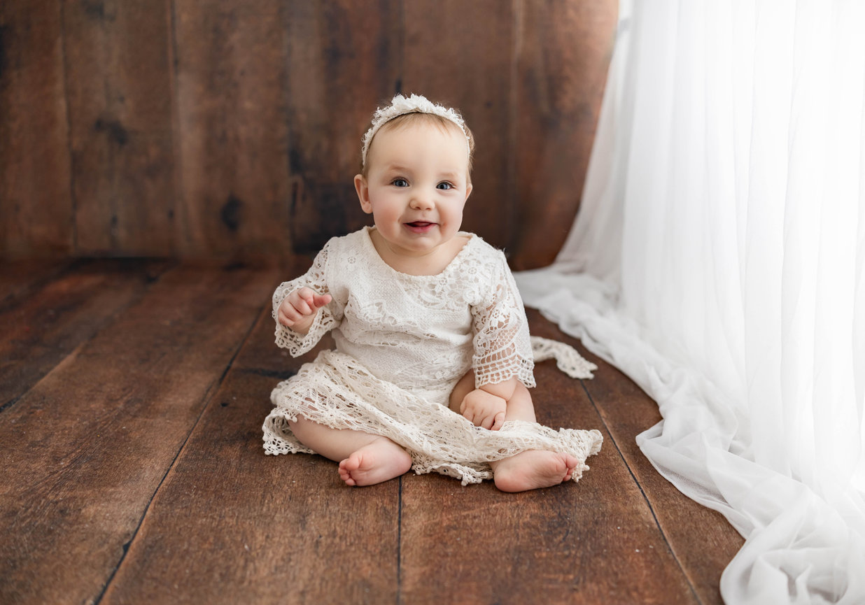 Baby sitting in cream dress against neutral indoor session background during 12-month milestone portrait holding number one