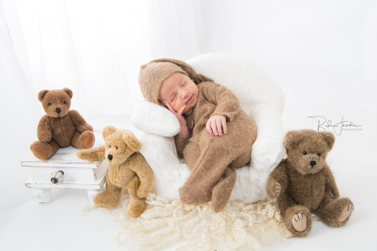 Newborn baby in a cozy outfit resting in a chair, surrounded by teddy bears. Soft colors and gentle ambiance.