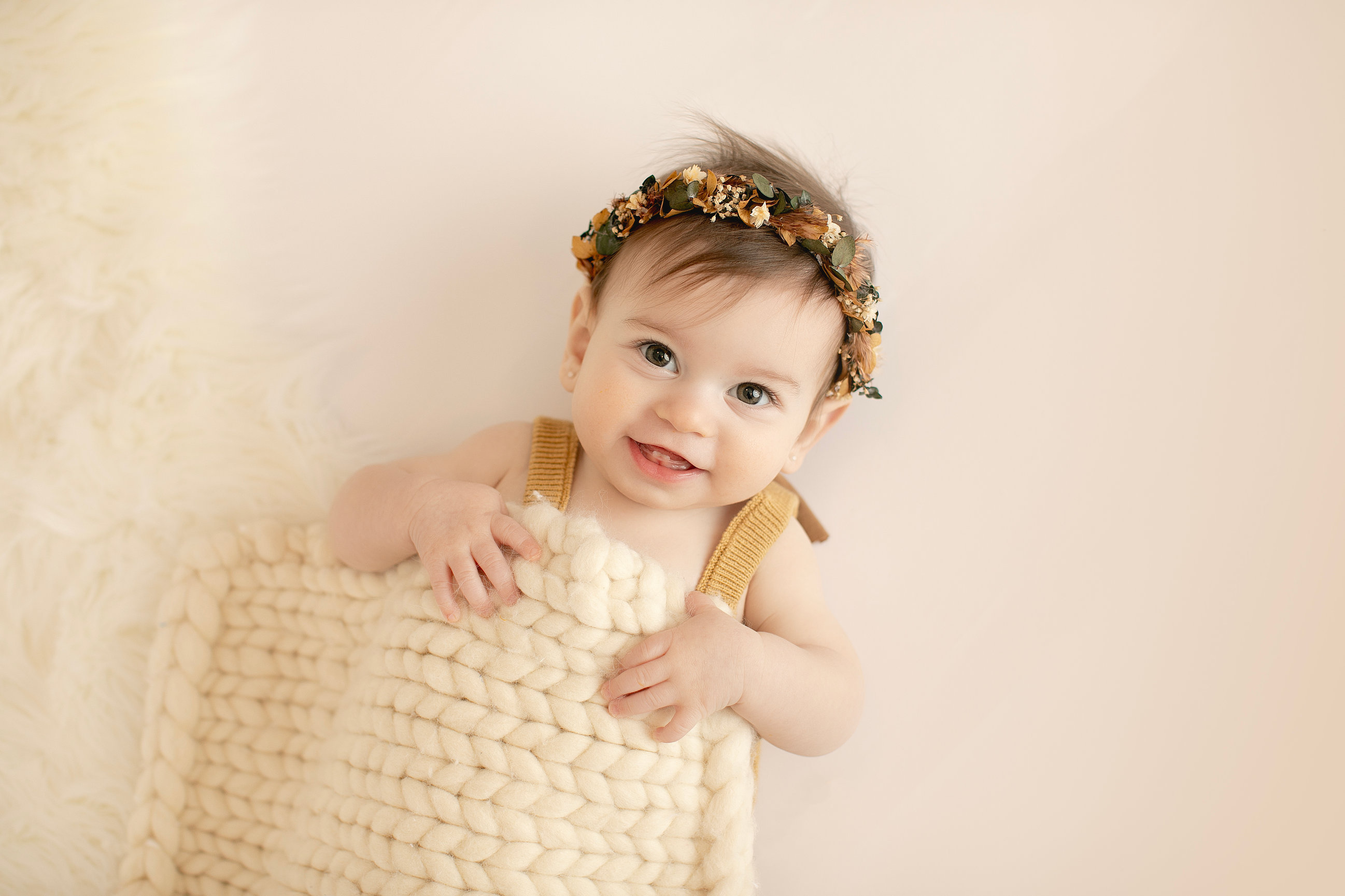 Baby in a mustard outfit peeking over a woven basket in a warm neutral studio set