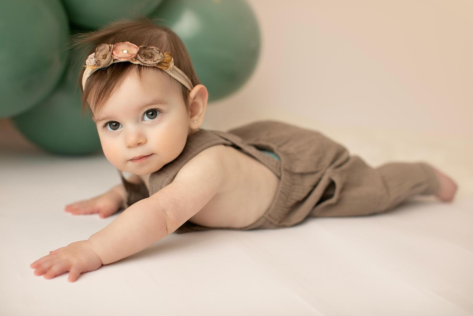 Baby stretching forward in a brown romper beside green balloons