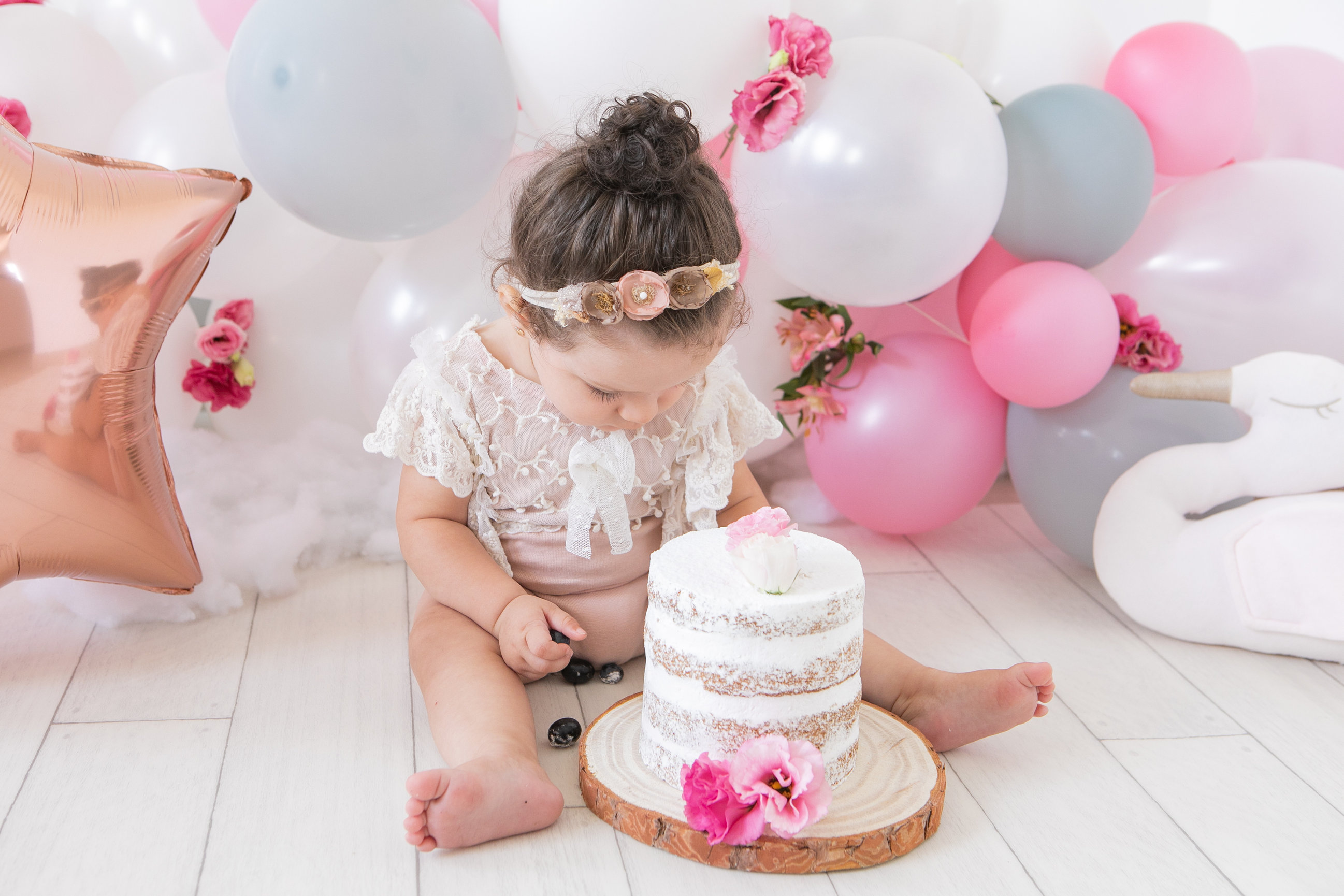 A toddler with a floral headband exploring a birthday cake among pink and white balloons.