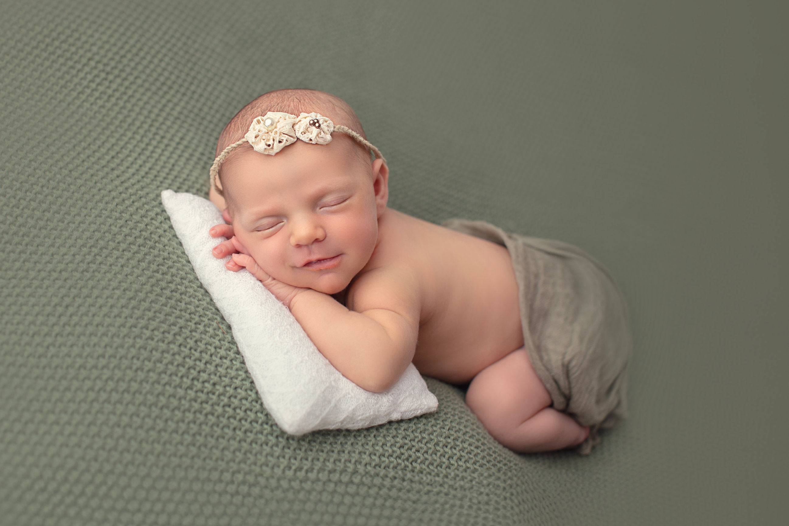 Sleeping newborn curled on sage fabric with a floral headband and tiny pillow.