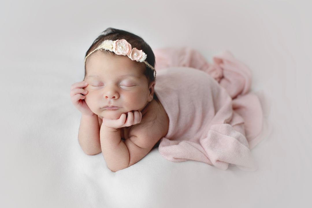 Sleeping newborn wearing a pink floral headband with hands beneath the chin.