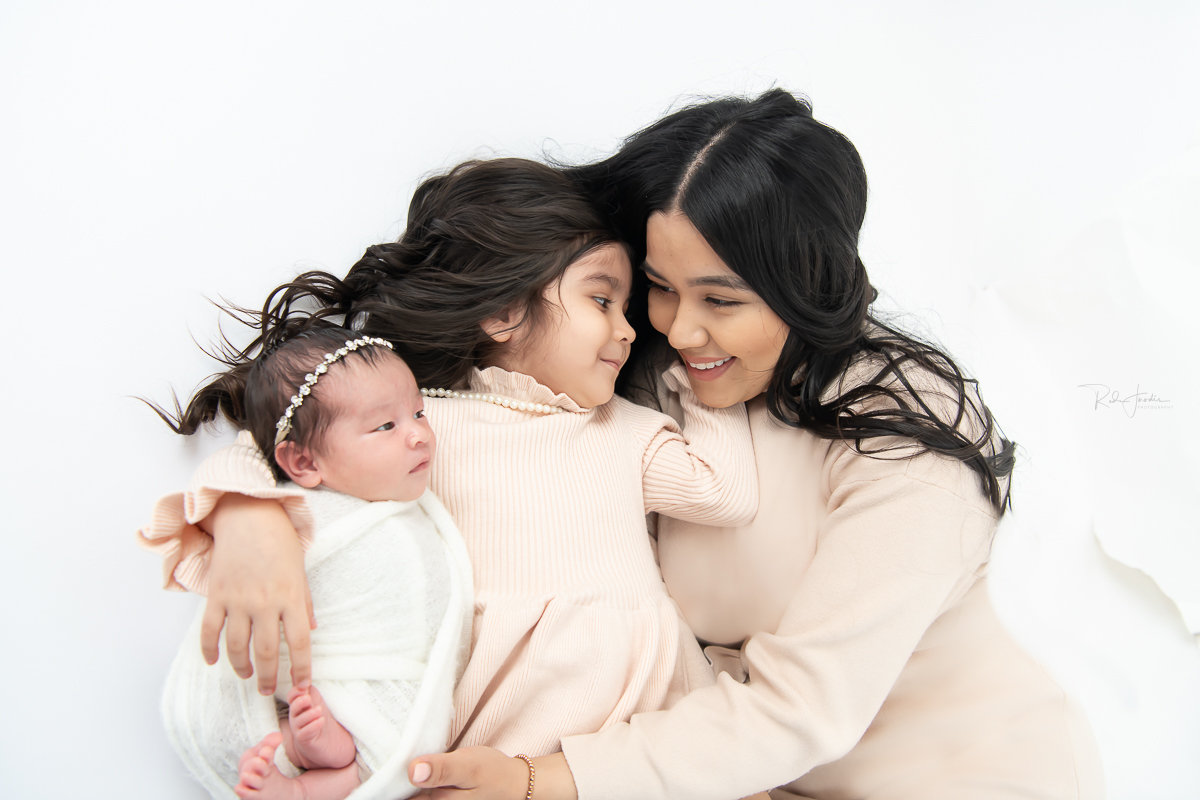Mother cuddles with her two daughters, a baby and a young girl, on a white background. Soft, warm atmosphere.