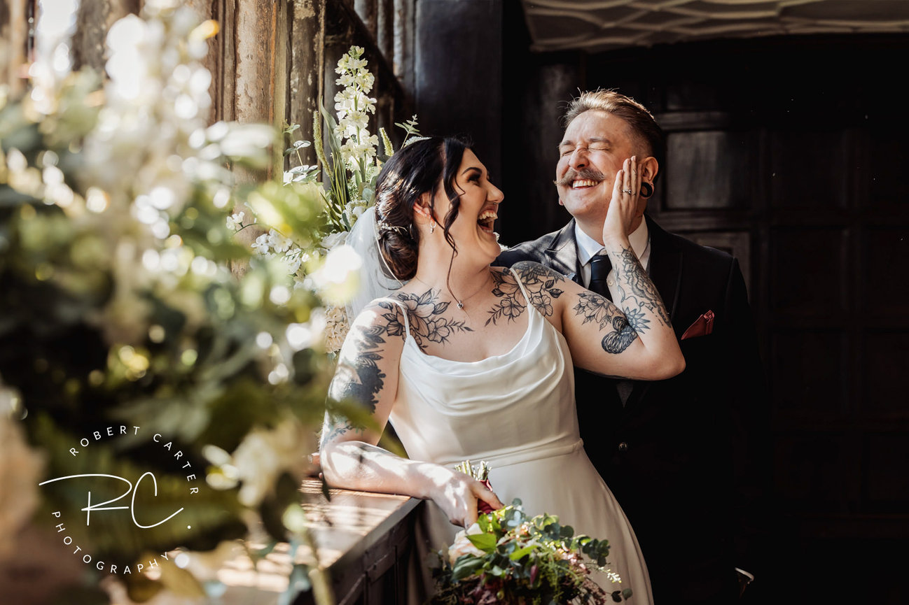 A joyful bride and groom share a laugh near a sunlit window, surrounded by greenery.