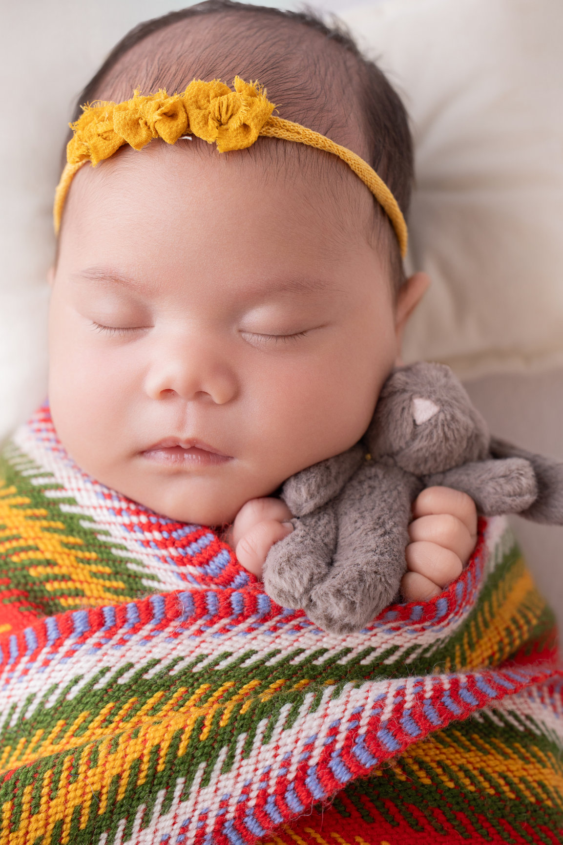 Sleeping newborn girl in a Métis sash holding a small grey teddy, mustard flower headband