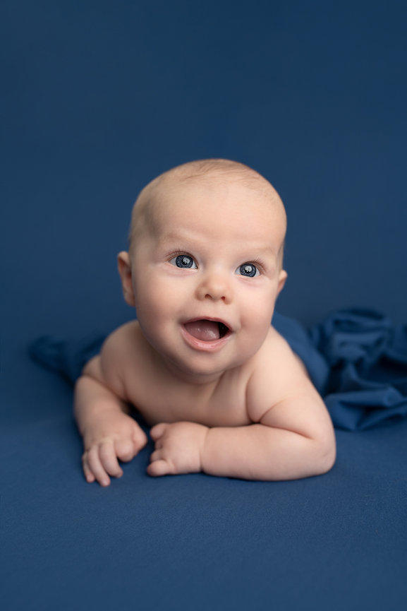 A baby with blonde hair is lying on their stomach on a blue surface, smiling and looking ahead. A soft, blue fabric partially covers the babys lower body. The background is also blue, perfect for capturing heartwarming baby photography near me.
