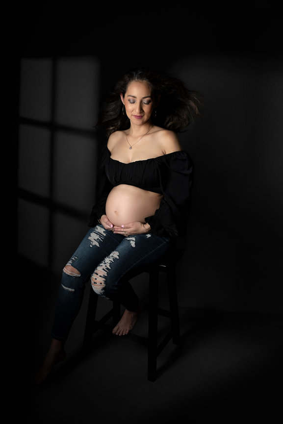 A pregnant woman with long, dark hair sits on a stool in a dark room. She wears a strapless black top and ripped jeans, holding her belly. Soft light falls on her from a window, casting a grid-like shadow on the floor and wall.