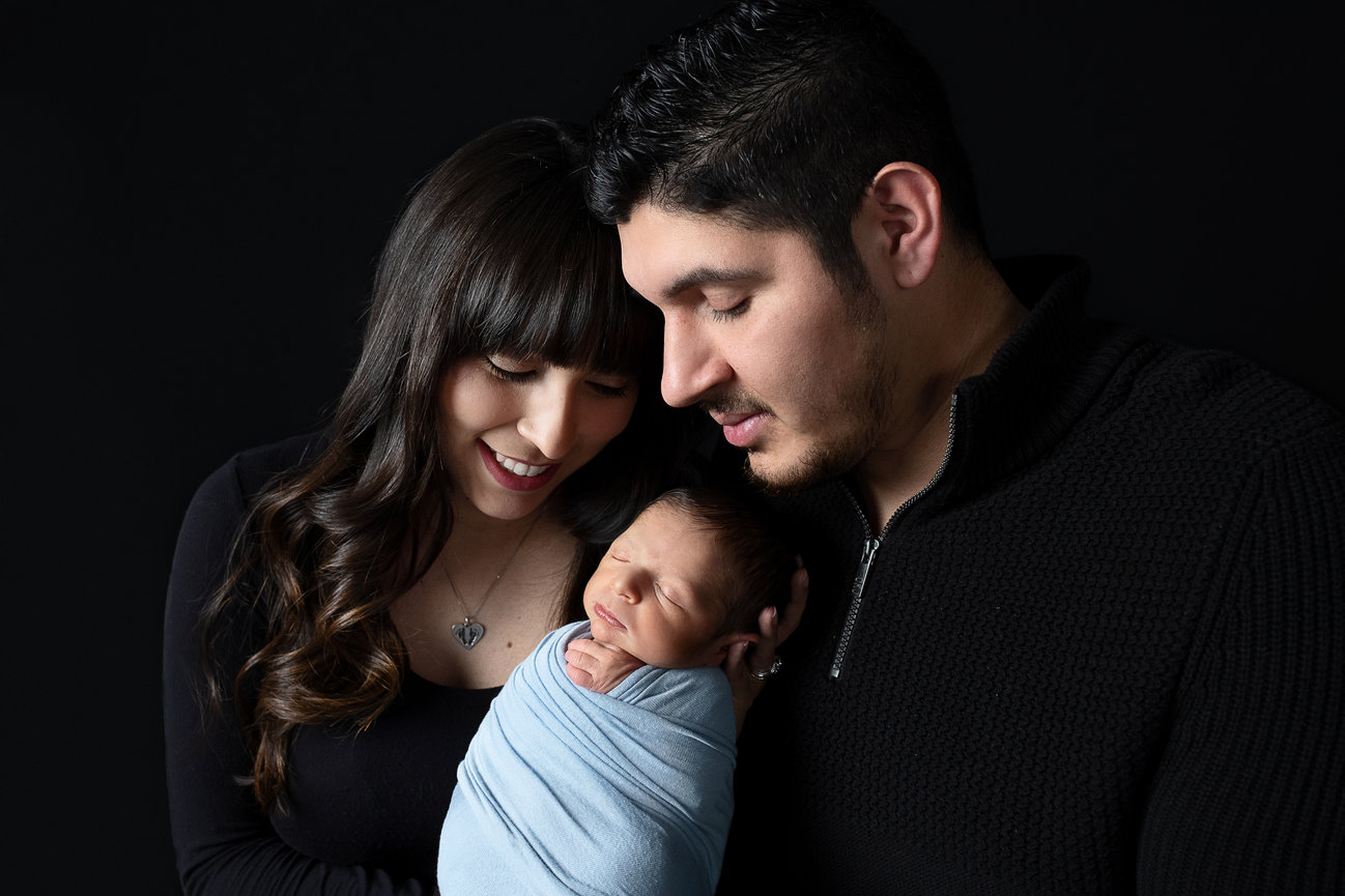 In Streamwood, Illinois, a couple lovingly gazes at their newborn wrapped in a soft blue blanket. The man and woman are wearing black tops and smile gently at the sleeping baby, set against a dark background.