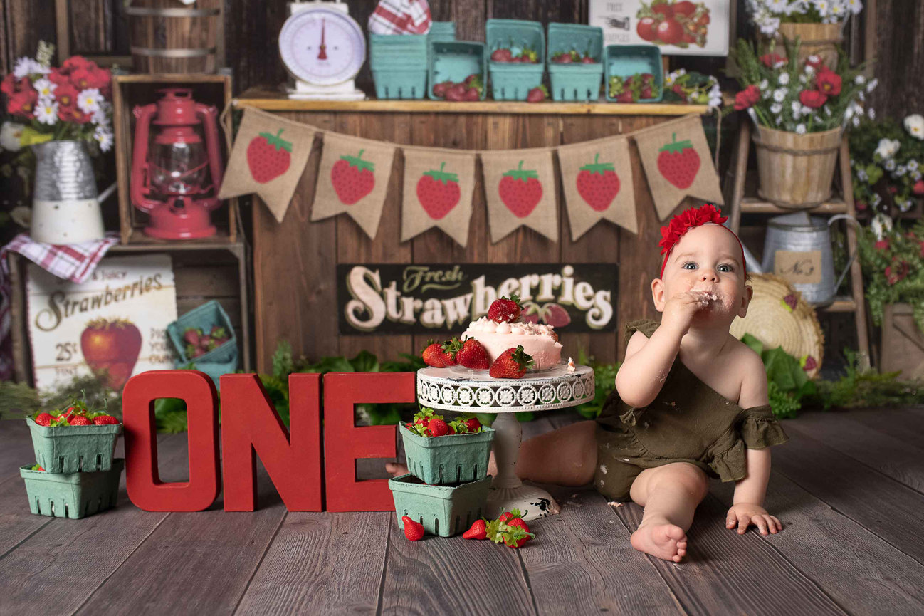 A baby celebrating their birthday sits beside a cake, wearing a red headband and eating with their hand. Surrounding are strawberry decor, baskets, red ONE letters, and a vintage berry stand backdrop adorned with festive signs featuring strawberries.