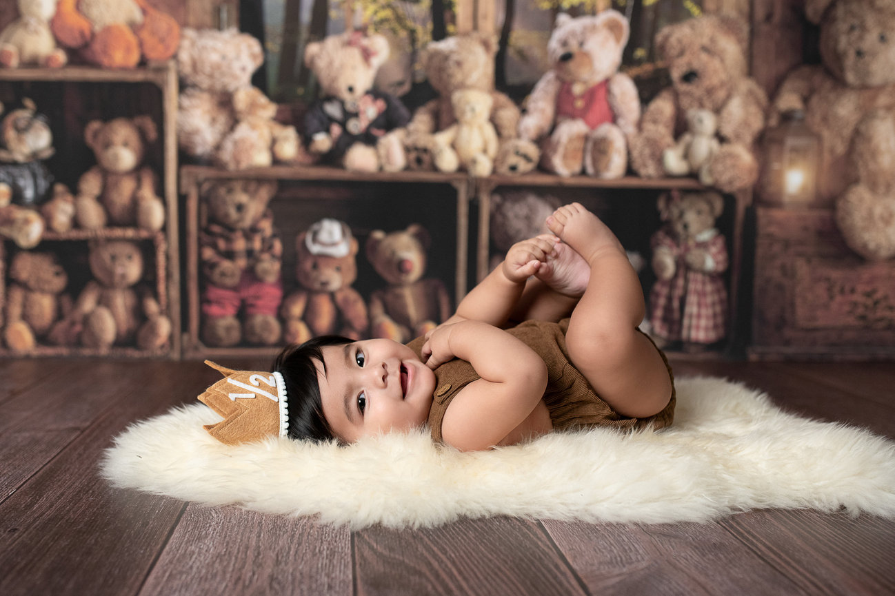 A six-month-old baby wearing a brown romper and a crown lies on a soft, fluffy rug. The backdrop is filled with shelves of teddy bears. The baby is smiling and holding their feet, creating a joyful and playful scene perfect for six-month photography.