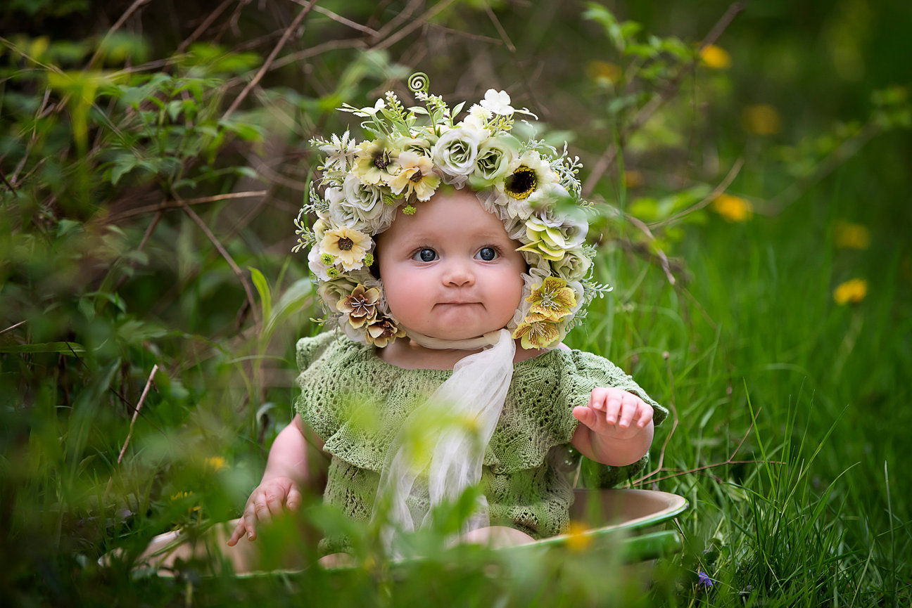 A baby sits in a lush, green garden surrounded by plants and flowers. Clad in a green knit outfit and a floral headband with blooms, they gaze at the camera, capturing the magic of the moment through the lens of a skilled 6-month photographer.