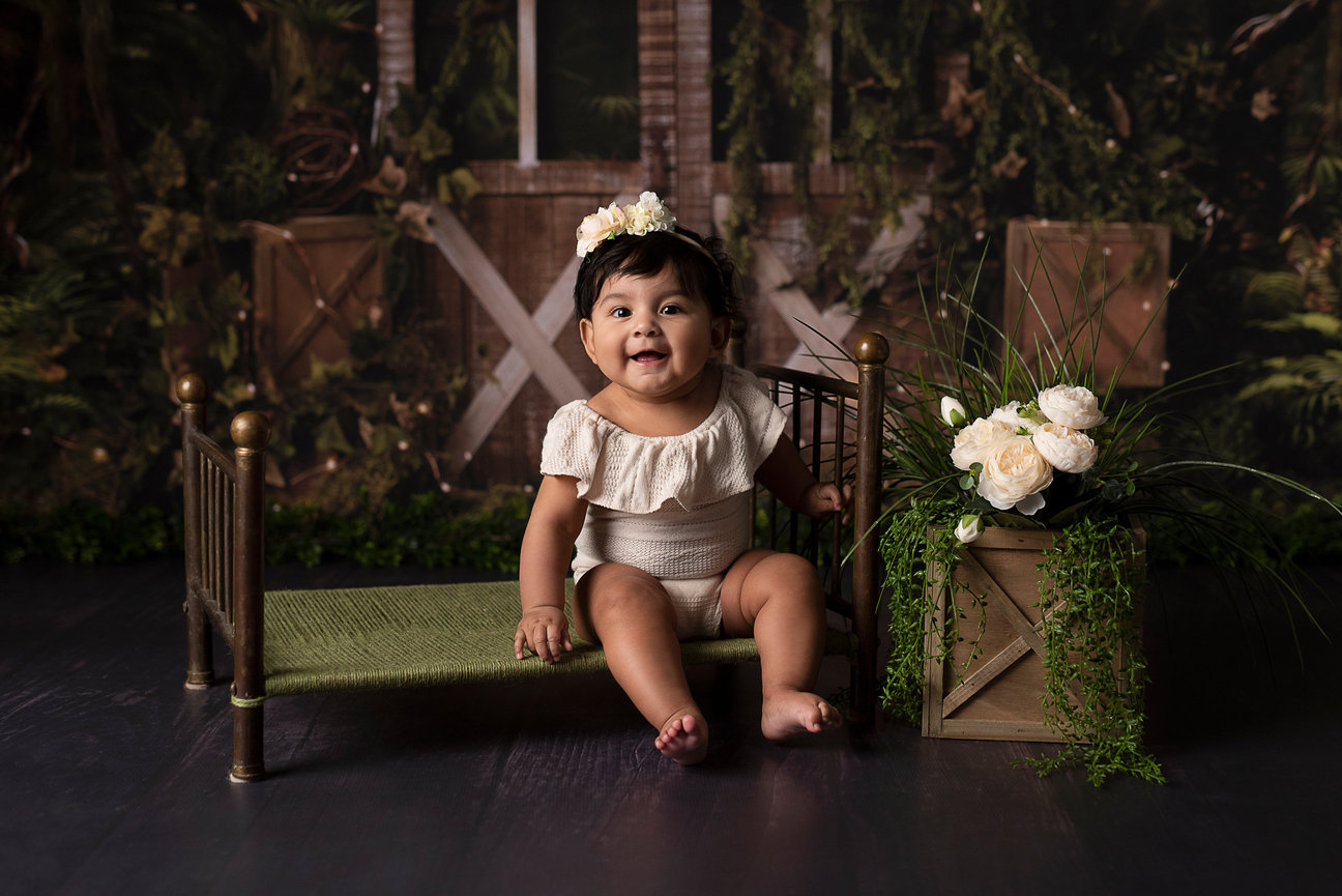 A baby sits on a small green bed, wearing a beige outfit and a floral headband in a photography studio near me. The backdrop is adorned with lush greenery and wooden elements, complemented by a crate with white flowers.