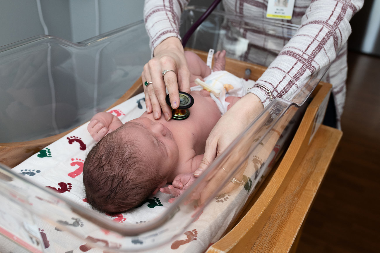 A newborn baby, nestled in a bassinet at Advocate Sherman Hospital, wears a white outfit, sleeping peacefully. Dark-haired with a tiny identification bracelet on their ankle, the infant is framed by a window view of the serene outdoor area beyond.
