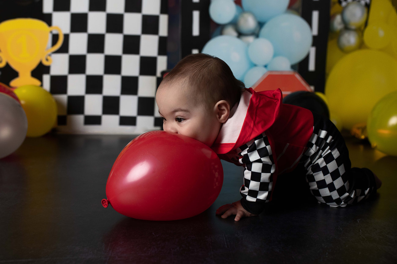 A baby in a checkered outfit leans on a red balloon with a race-themed background.