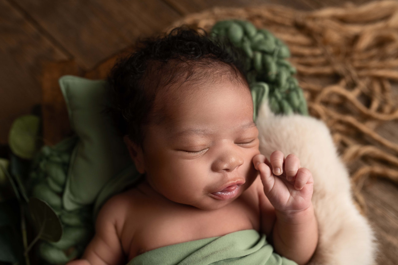 A sleeping baby wrapped in a green blanket lies on a fluffy surface. The baby has curly hair and is resting peacefully, surrounded by soft textures in a cozy setting.