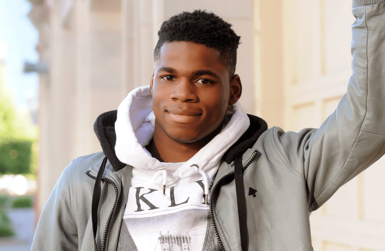 An African American young man in a graphic hoodie stands against a brick wall.