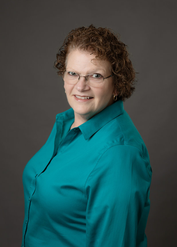 A woman with curly hair and glasses smiles, wearing a teal shirt against a gray background.