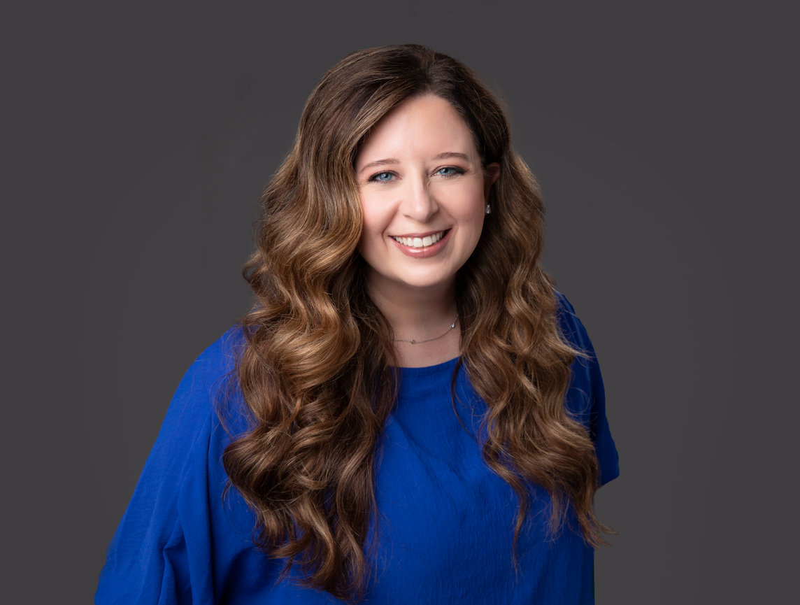Smiling woman with long, wavy hair wearing a blue shirt against a dark background.
