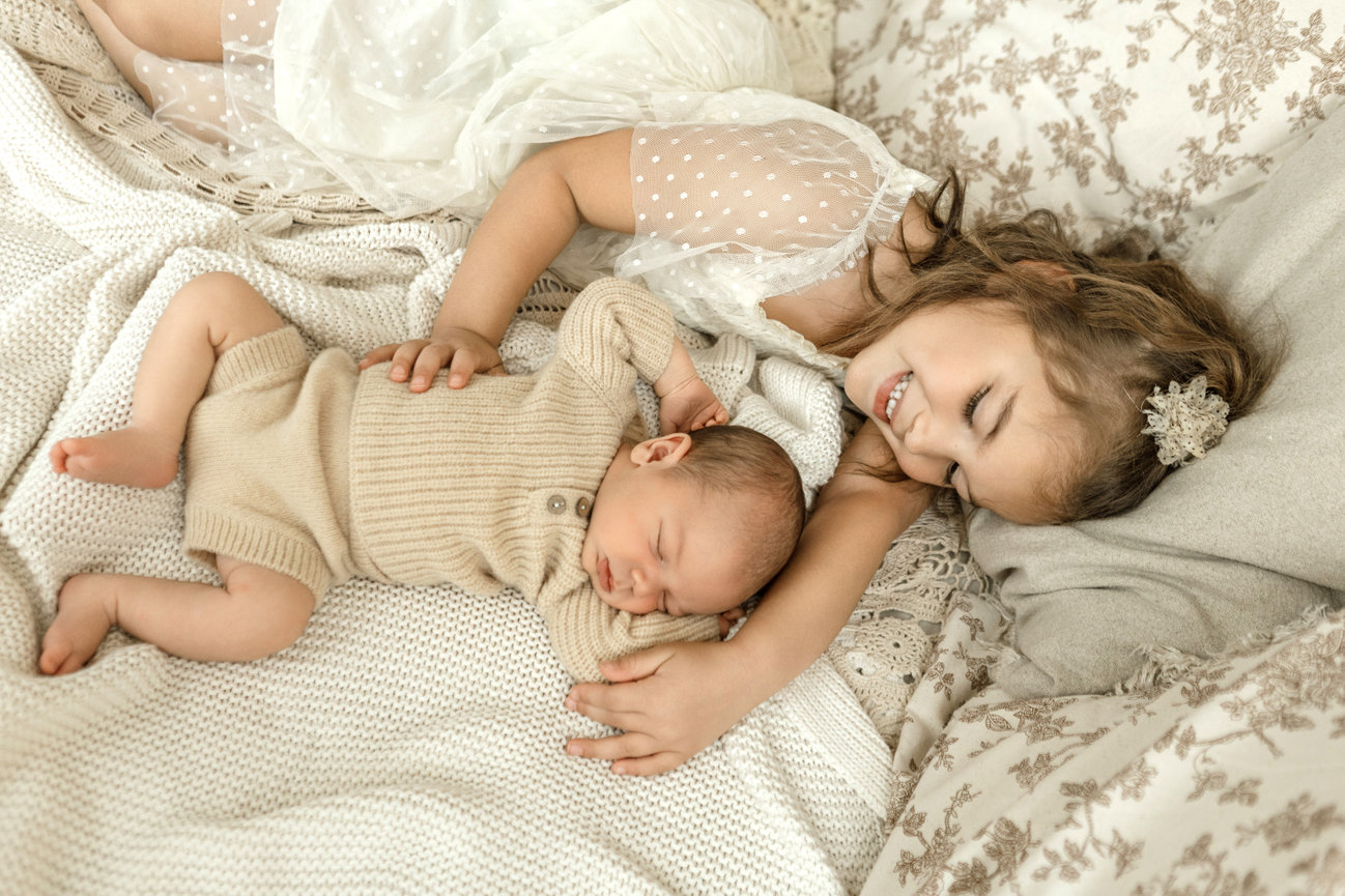 A smiling girl lies next to a peacefully sleeping baby, both on a cozy, textured blanket.