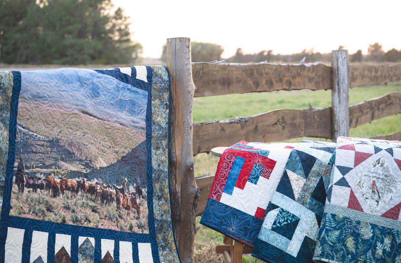 Colorful quilts displayed against a rustic wooden fence with a grassy field in the background.