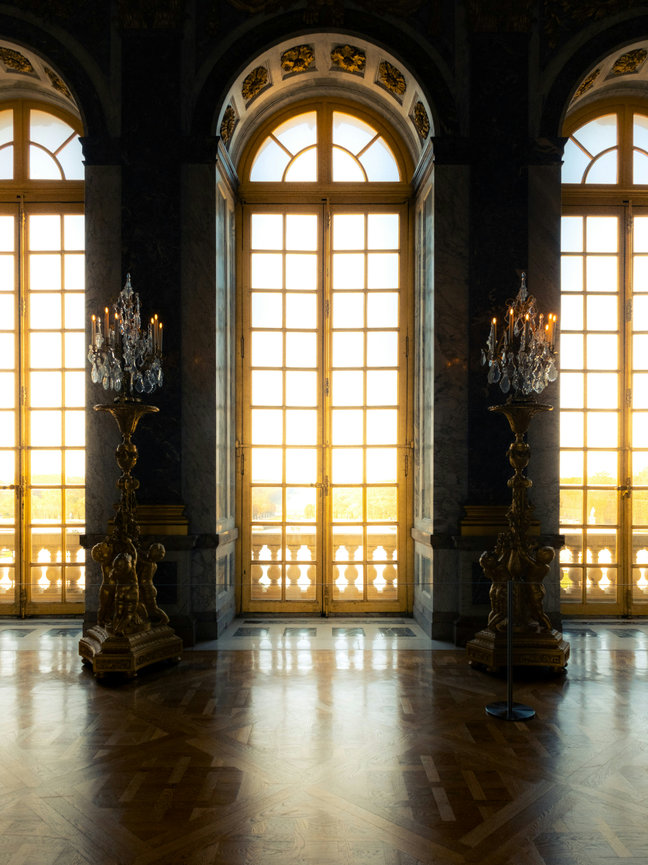 Elegant room with tall arched windows, ornate chandeliers, and polished wooden floor, illuminated by warm natural light.