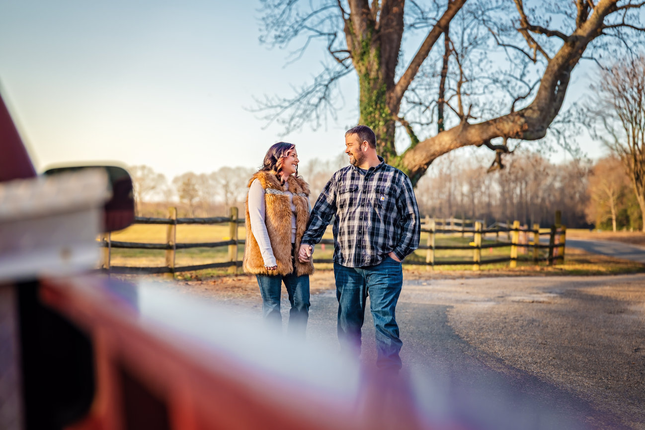 Outdoor photography session of a couple holding hands, walking by a wooden fence in a rural setting with bare trees and clear sky.