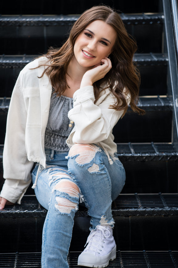 Teen girl sitting on concrete stairs in a gritty urban setting, wearing distressed jeans and a denim jacket.