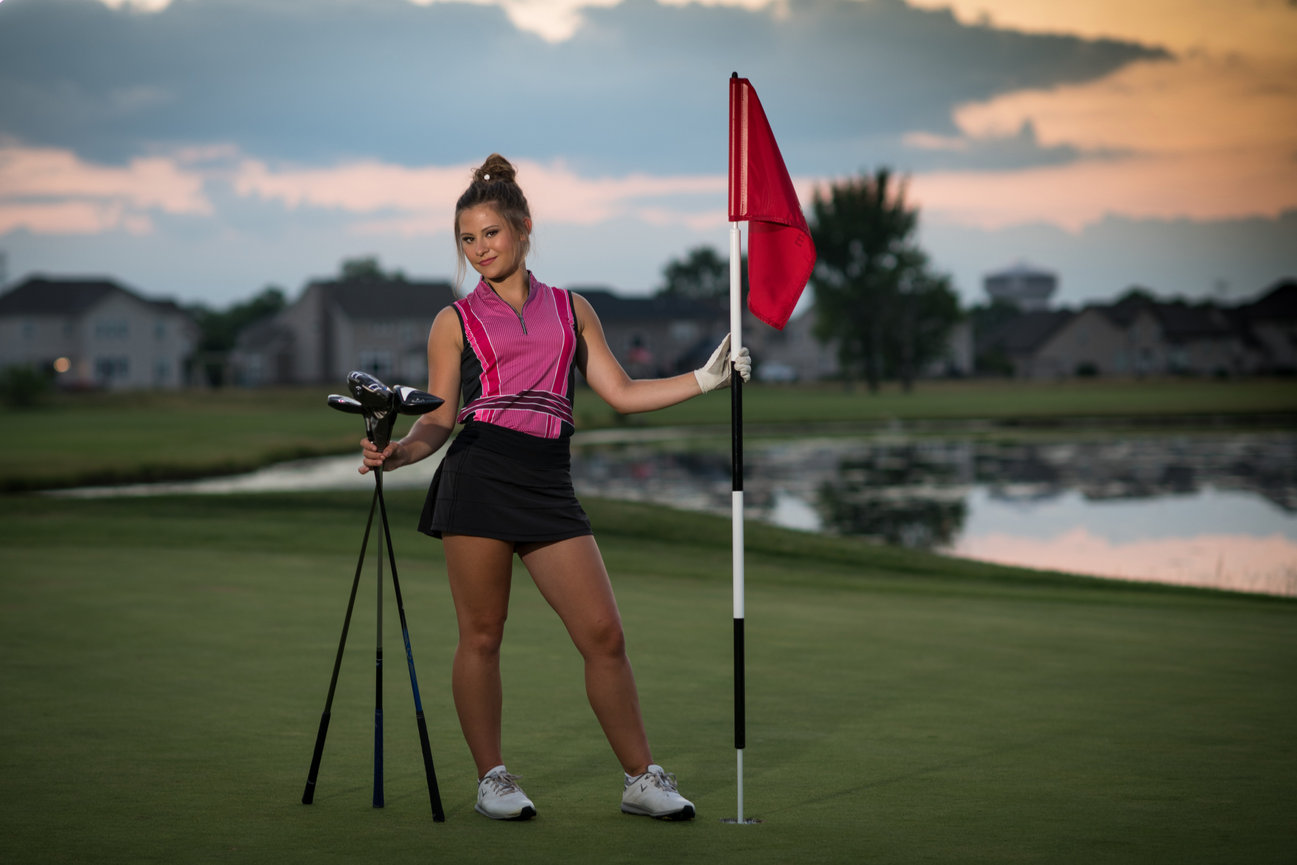 Golfer in a stylish outfit poses on the green at sunset, holding clubs and a flag.