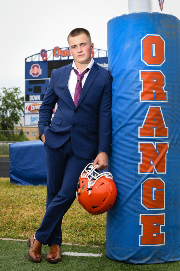 A young man in a suit leans against a football goalpost, holding an orange helmet, on a high school field.
