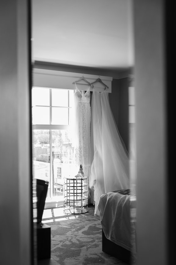 Wedding dress and veil hanging in a bright hotel room, with a view through a doorway.