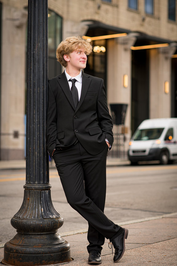 Teen guy in a black suit leans on a lampost, smiling, in a downtown urban environment for senior pictures in Columbus Ohio.