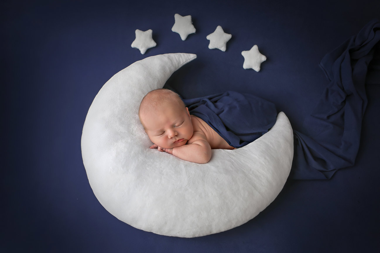 Baby sleeping on a crescent moon-shaped pillow with stars above on a dark blue background.