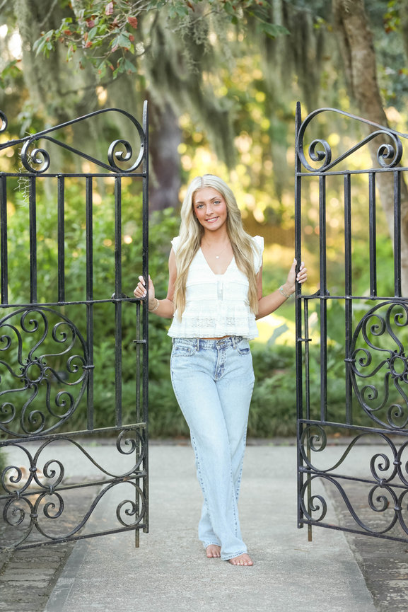 A woman in a white top and jeans stands barefoot between ornate iron gates in a lush garden setting.