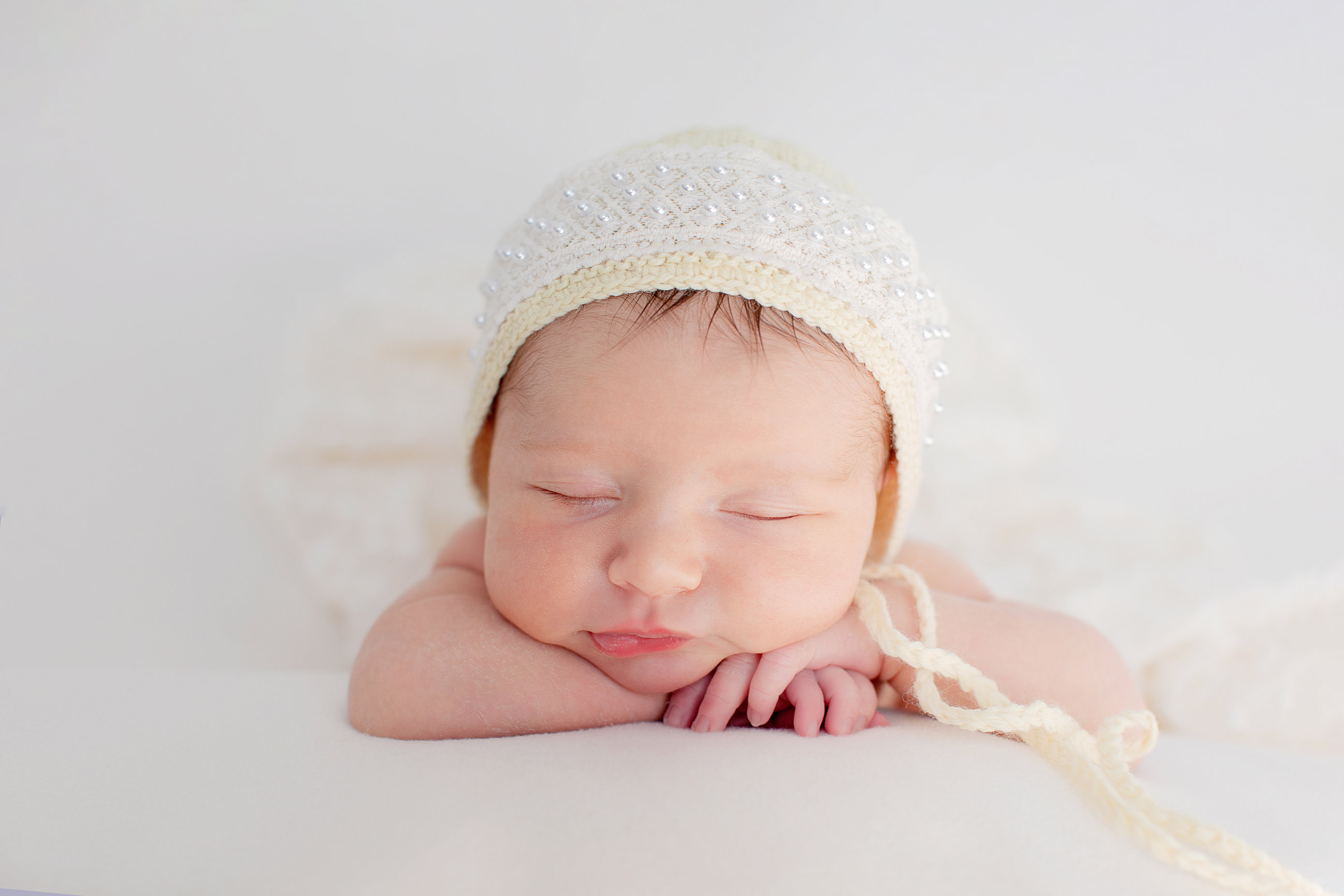 Sleeping newborn wearing a cream knitted bonnet, resting on folded hands.