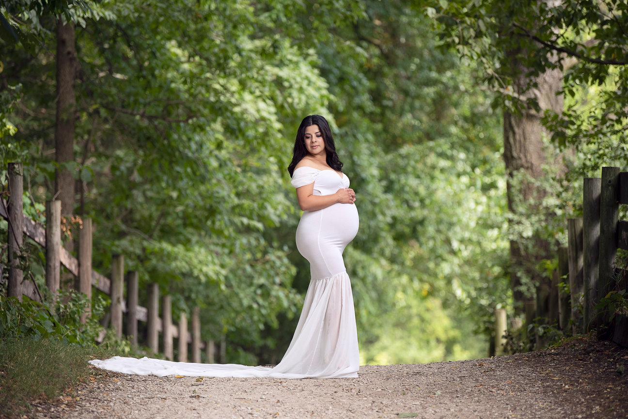 A pregnant woman in a long, flowing white dress stands on a wooded path. She is gently holding her belly and surrounded by lush green trees. The sunlight filters softly through the leaves.