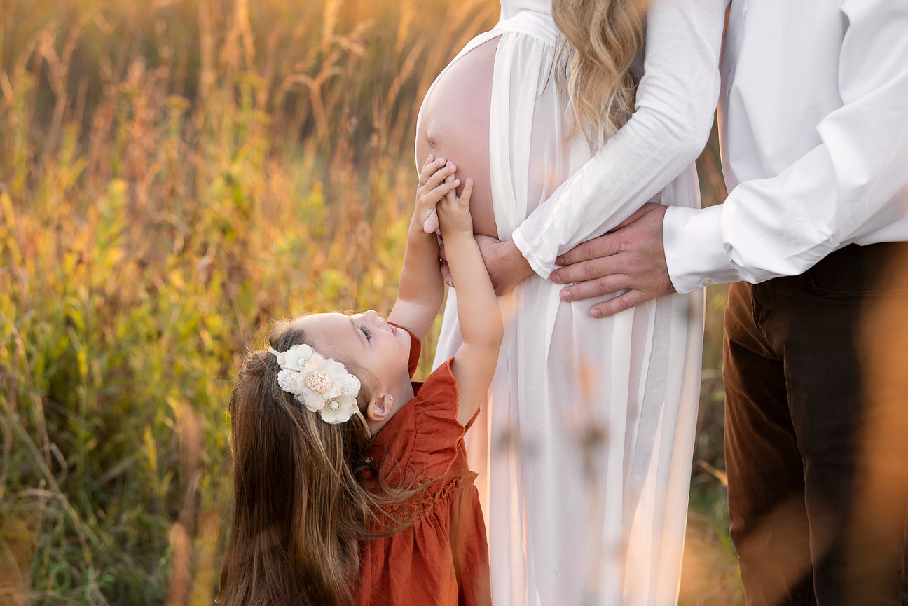 A little girl in a red dress gently touches a pregnant womans belly in a grassy field. The woman is wearing a white dress, and an adult stands beside them. The scene is warm and serene, highlighting a caring family moment.